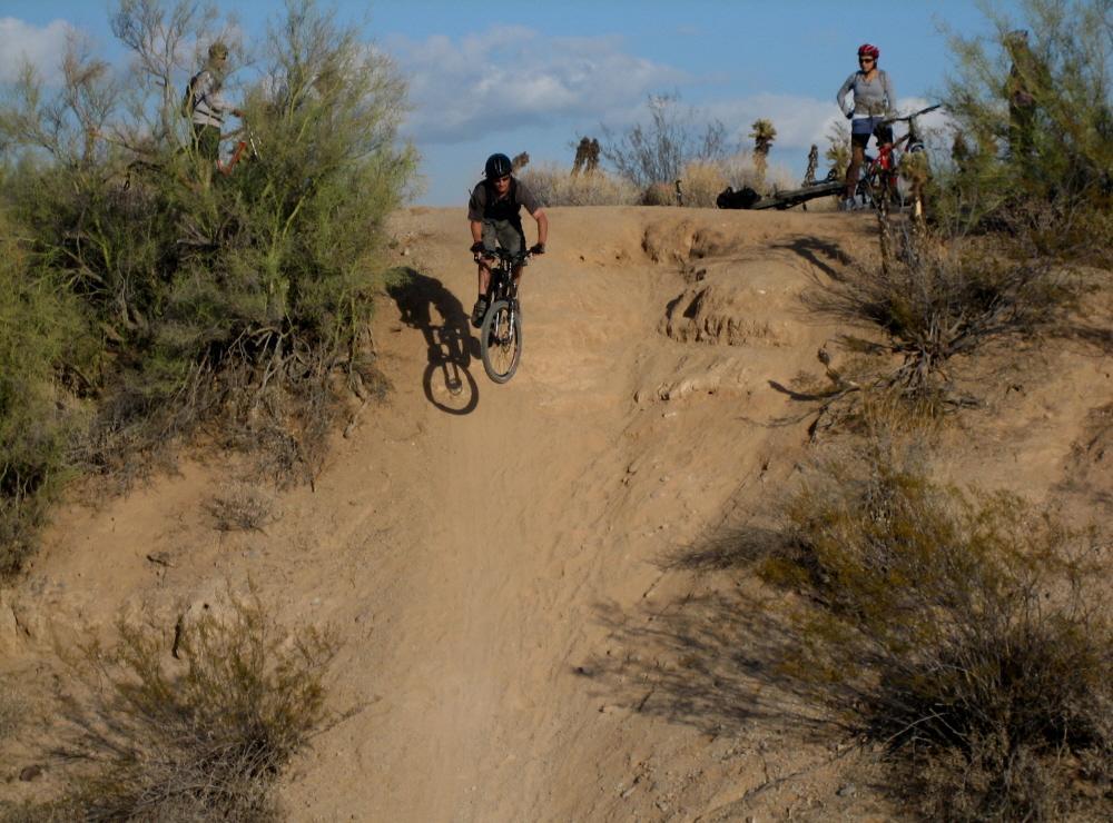 A mountain biker in mid-air, jumping off a dirt ramp in a desert landscape, with two other cyclists on the trail nearby, surrounded by sparse vegetation and rocky terrain under a blue sky with clouds. McDowell Mountain Park mountain bike trail.