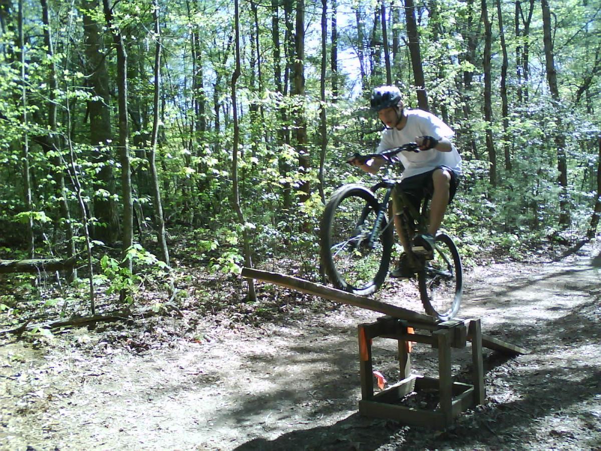 A person riding a mountain bike performs a jump over a wooden plank set up in a forested area, surrounded by lush green trees and sunlight filtering through the leaves. Dark Mountain Trail mountain bike trail.