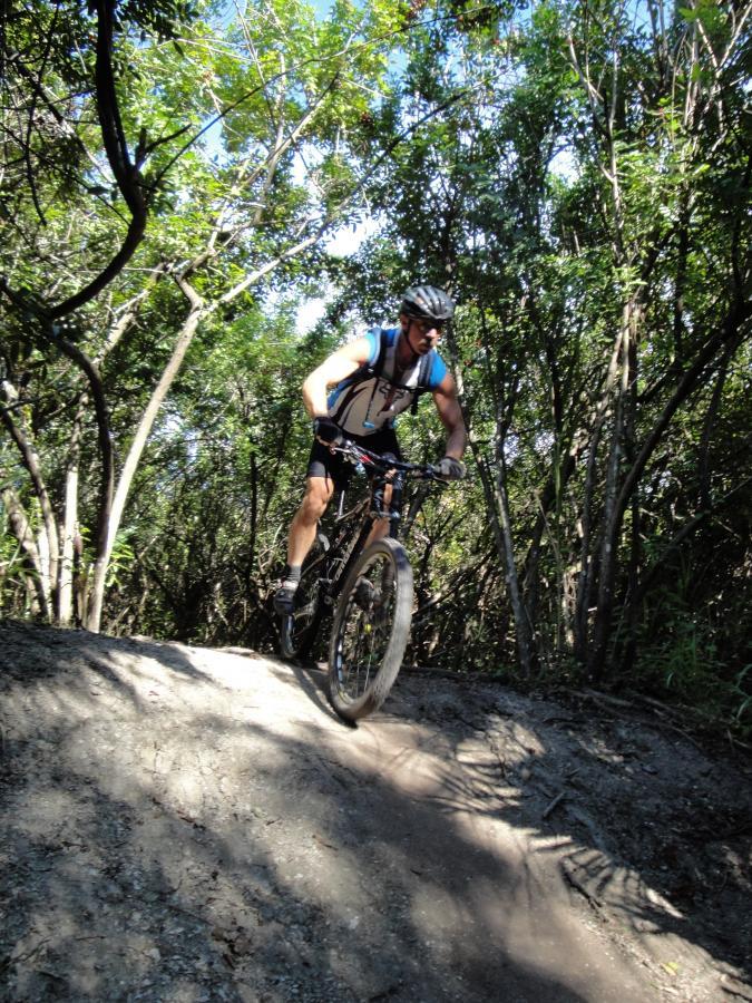 A mountain biker navigating a dirt trail in a wooded area, with sunlight filtering through the trees. Caloosahatchee Regional Park mountain bike trail.