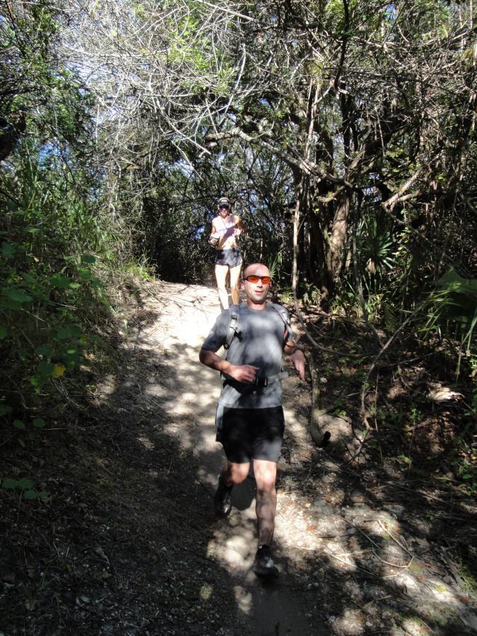 A man running on a dirt path through a wooded area, wearing a gray shirt and sunglasses. In the background, a woman is walking, also in athletic attire. Sunlight filters through the trees, casting shadows on the path. Caloosahatchee Regional Park mountain bike trail.