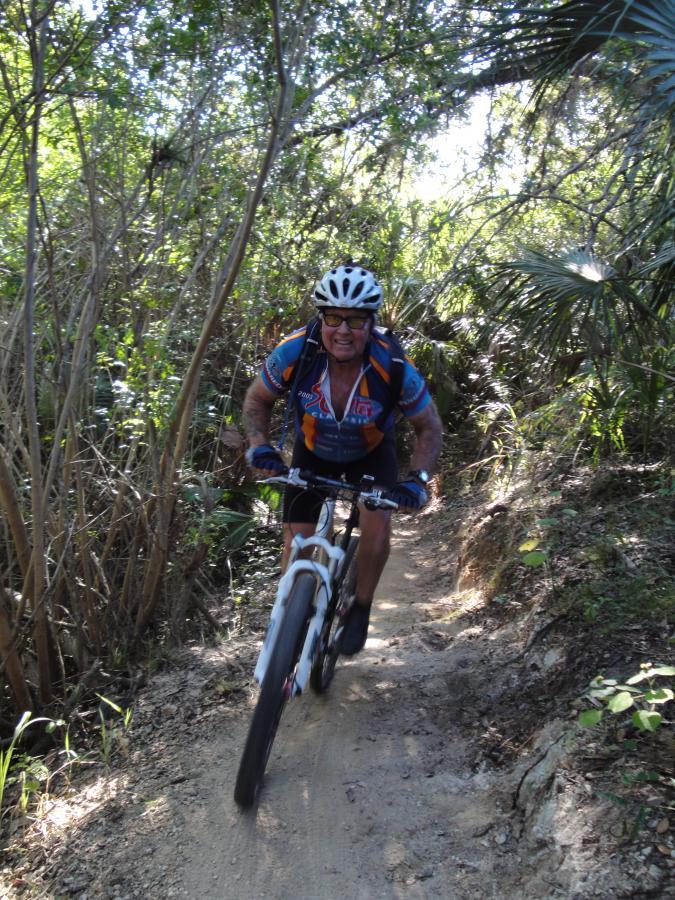 A mountain biker in a blue and orange jersey rides uphill on a narrow dirt trail surrounded by dense foliage and sunlight filtering through the trees. He is wearing a helmet and sunglasses, focused on navigating the challenging terrain. Caloosahatchee Regional Park mountain bike trail.