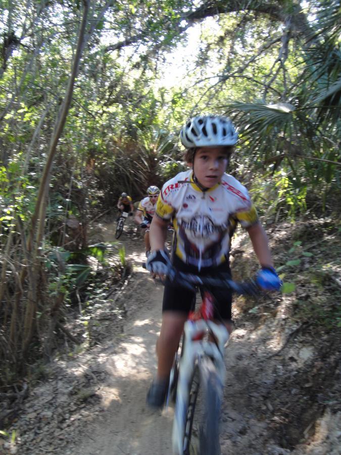 A group of young mountain bikers riding on a narrow dirt trail surrounded by dense greenery. The focus is on one cyclist in the foreground, wearing a helmet and sports attire, while others are visible in the background. The scene captures the excitement of a mountain biking adventure in a natural setting. Caloosahatchee Regional Park mountain bike trail.