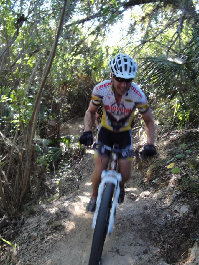 A cyclist in a helmet and sportswear maneuvers through a narrow, wooded trail on a mountain bike, showing determination as they navigate the terrain. The surrounding greenery includes dense foliage and trees typical of a forested area. Caloosahatchee Regional Park mountain bike trail.