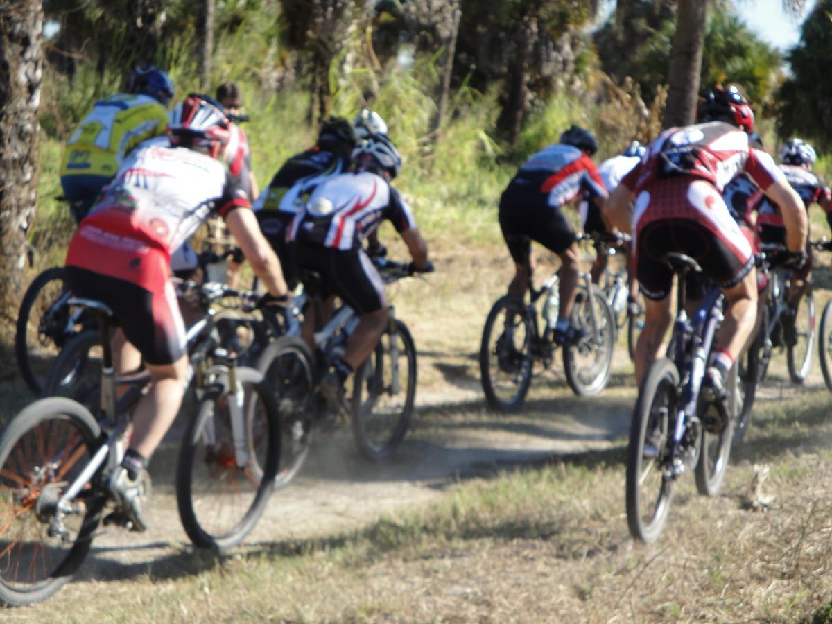 A group of mountain bikers in athletic gear riding on a dirt trail, surrounded by greenery. The image captures the cyclists from behind, showcasing their dynamic movement and the dust being kicked up from the trail. Caloosahatchee Regional Park mountain bike trail.