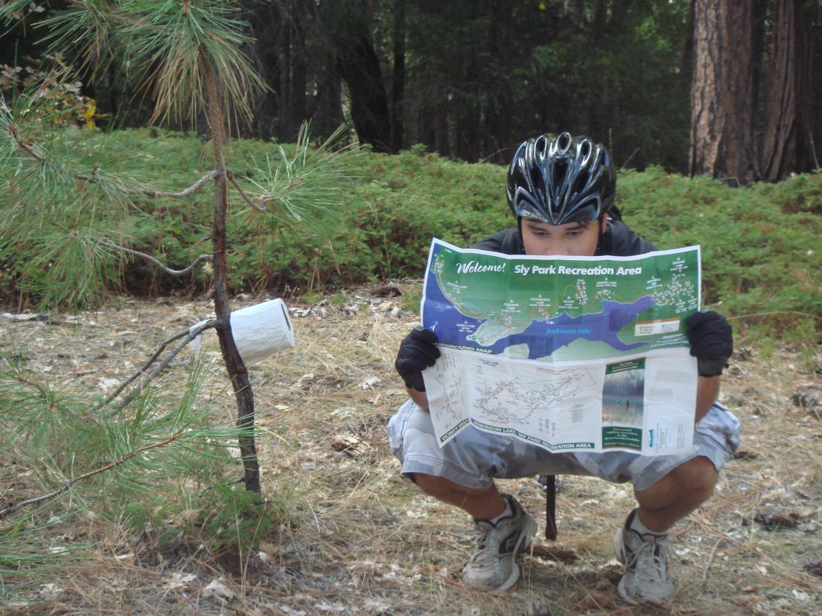 A person squatting in a forested area, wearing a helmet and gloves, studying a map labeled "Sly Park Recreation Area." Nearby, a small white container is attached to a tree. The ground is covered with pine needles, and there are lush green plants in the background. Jenkinson Lake Loop mountain bike trail.