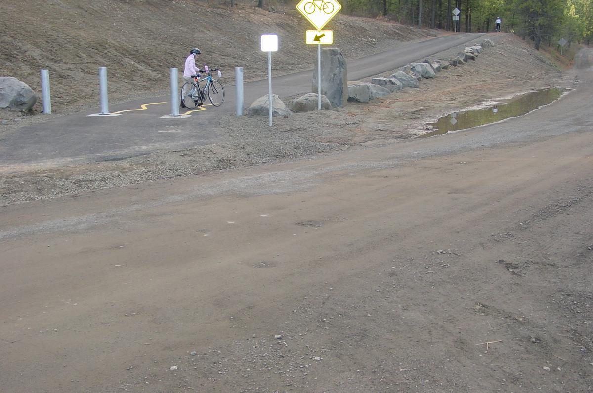 A cyclist in a light pink jacket and helmet is walking their bike along a dirt path. In the background, a curving road is visible, bordered by rocks. Signs indicate bicycle direction and caution. The area appears to be forested, with sparse trees lining the path. A small puddle is present on the right side of the image. Fish Lake Trail mountain bike trail.