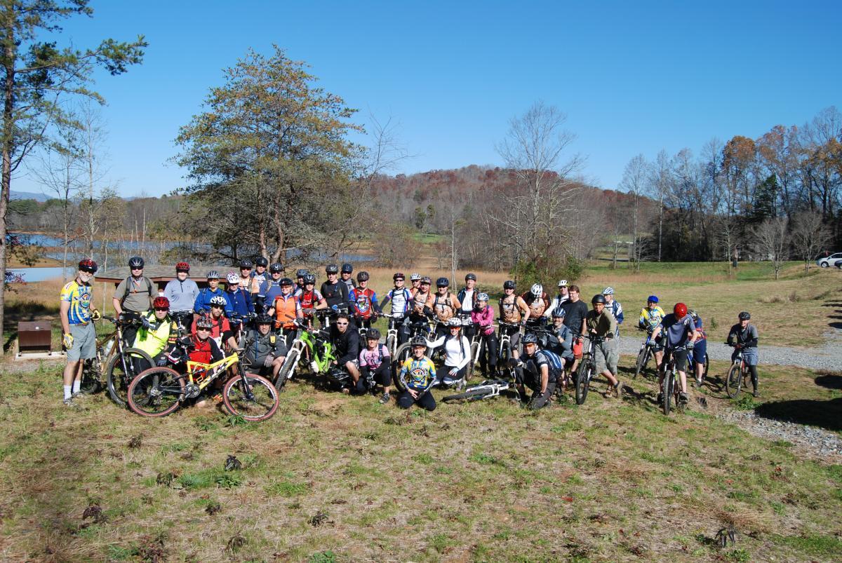 A diverse group of over 40 mountain bikers posing together on a grassy area near a lake, with trees and hills in the background. They are wearing helmets and colorful biking gear, some holding their bikes while others sit or kneel on the ground. The sky is clear and blue, indicating a bright and sunny day. Jack Rabbit Trails mountain bike trail.