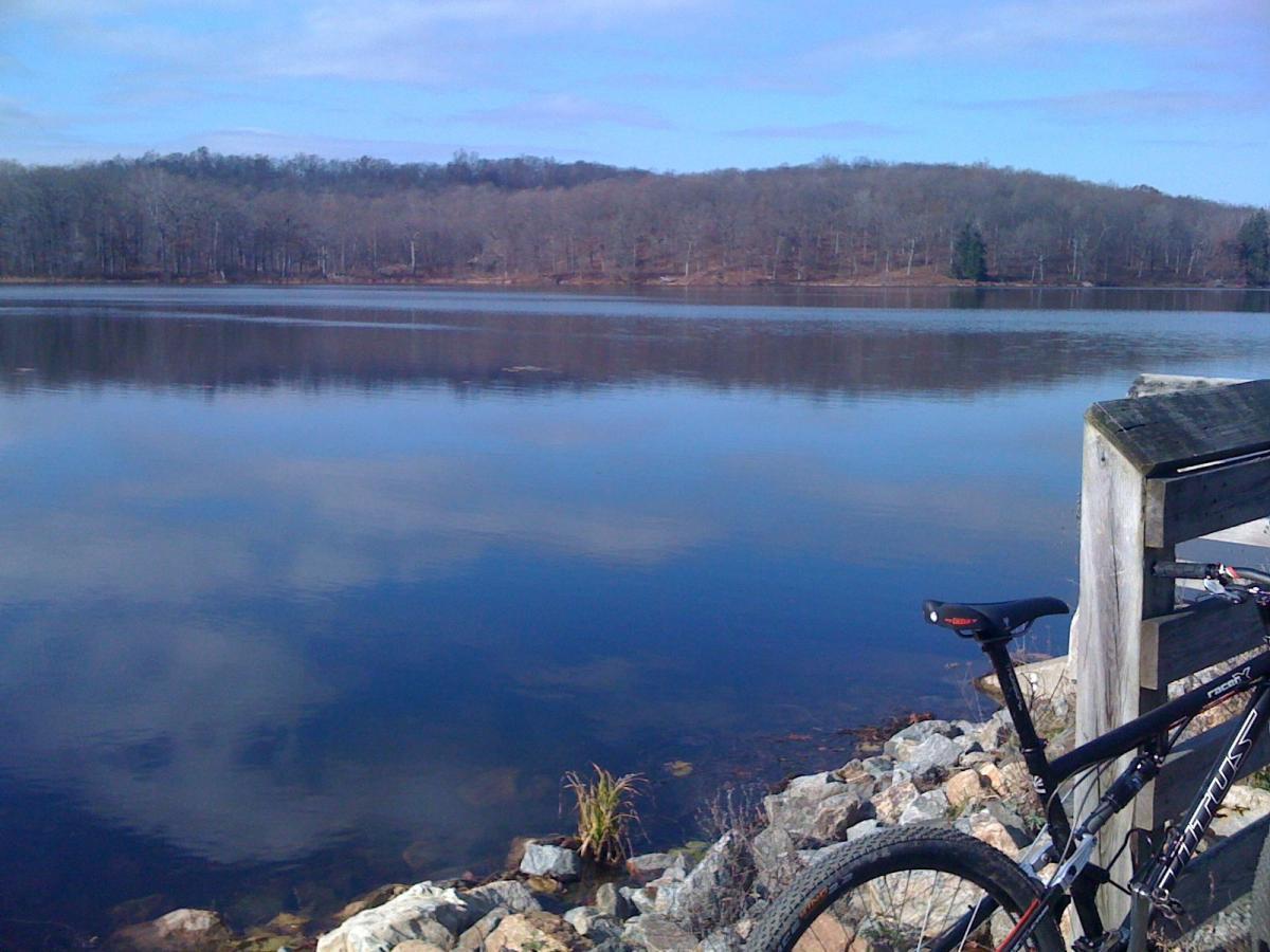 A serene landscape featuring a calm lake reflecting the sky, surrounded by bare trees on a distant hillside. In the foreground, there are smooth rocks and a bicycle resting against a wooden railing, suggesting a peaceful outdoor setting ideal for relaxation or biking. Allamuchy Mt. State Park: Deer Park mountain bike trail.