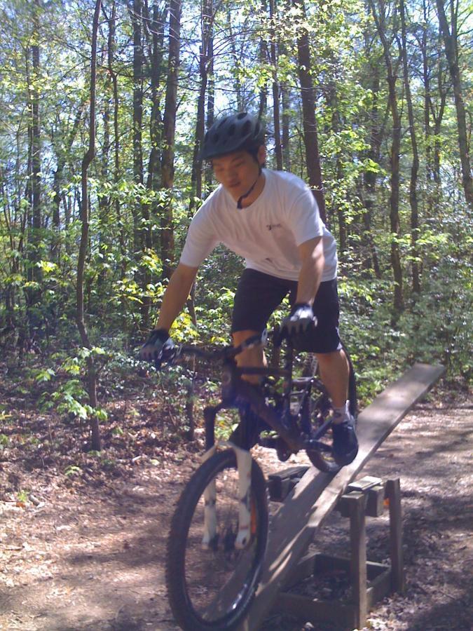 A person riding a mountain bike on a wooden ramp in a forested area. The rider is wearing a helmet and casual clothing, focused on balancing while navigating the obstacle amid greenery and trees. Dark Mountain Trail mountain bike trail.