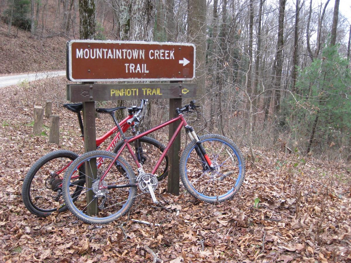 Alt text: A wooden trail sign labeled "Mountaintown Creek Trail" and "Pinhoti Trail" stands near two parked mountain bikes on a forested path covered with fallen leaves. The surroundings feature bare trees and a winding gravel road in the background. Pinhoti Trail: Mountaintown Creek Segment mountain bike trail.