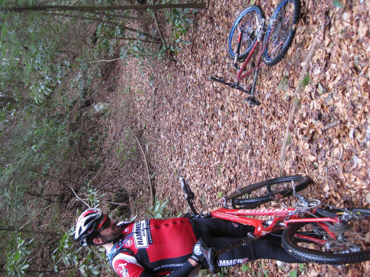 A mountain biker in a red and black cycling jersey stands next to a fallen bicycle on a leaf-covered trail in a forest. Surrounding vegetation includes trees and shrubs, indicating a natural setting conducive to outdoor biking activities. The cyclist is wearing a helmet and appears to be taking a break or assessing the situation. Pinhoti Trail: Mountaintown Creek Segment mountain bike trail.