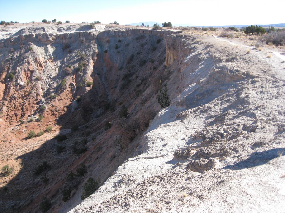 A panoramic view of a rugged canyon landscape with steep, rocky cliffs and sparse vegetation. The foreground shows a narrow, winding path along the edge of the canyon, while the background features layers of red and gray rock formations under a clear blue sky. White Ridge Bike Trails mountain bike trail.