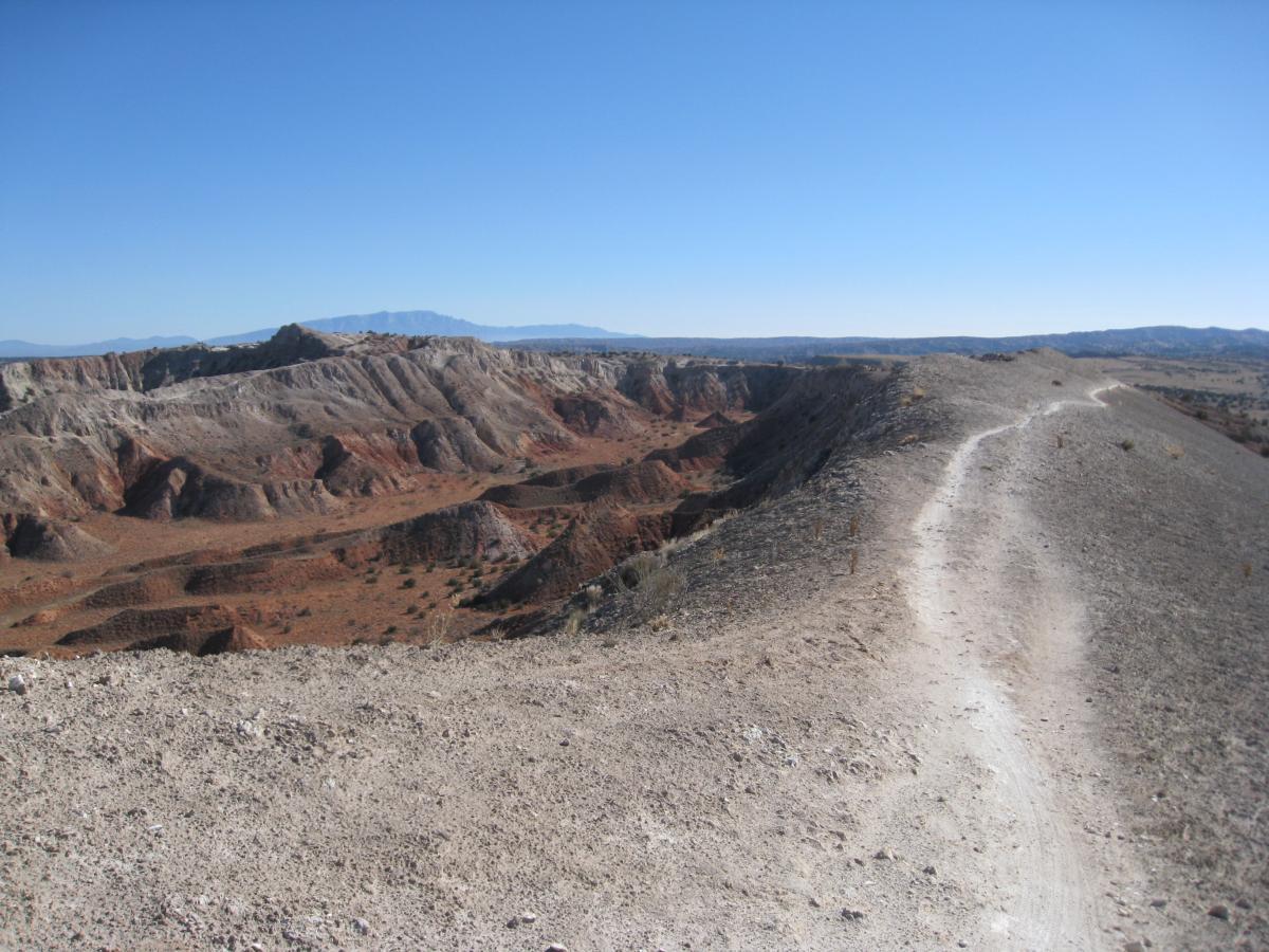 A panoramic view of a rugged landscape featuring layered rock formations in shades of red and gray, with a dirt trail winding along the crest of a hill. The sky above is clear and blue, and distant mountains are visible on the horizon. White Ridge Bike Trails mountain bike trail.