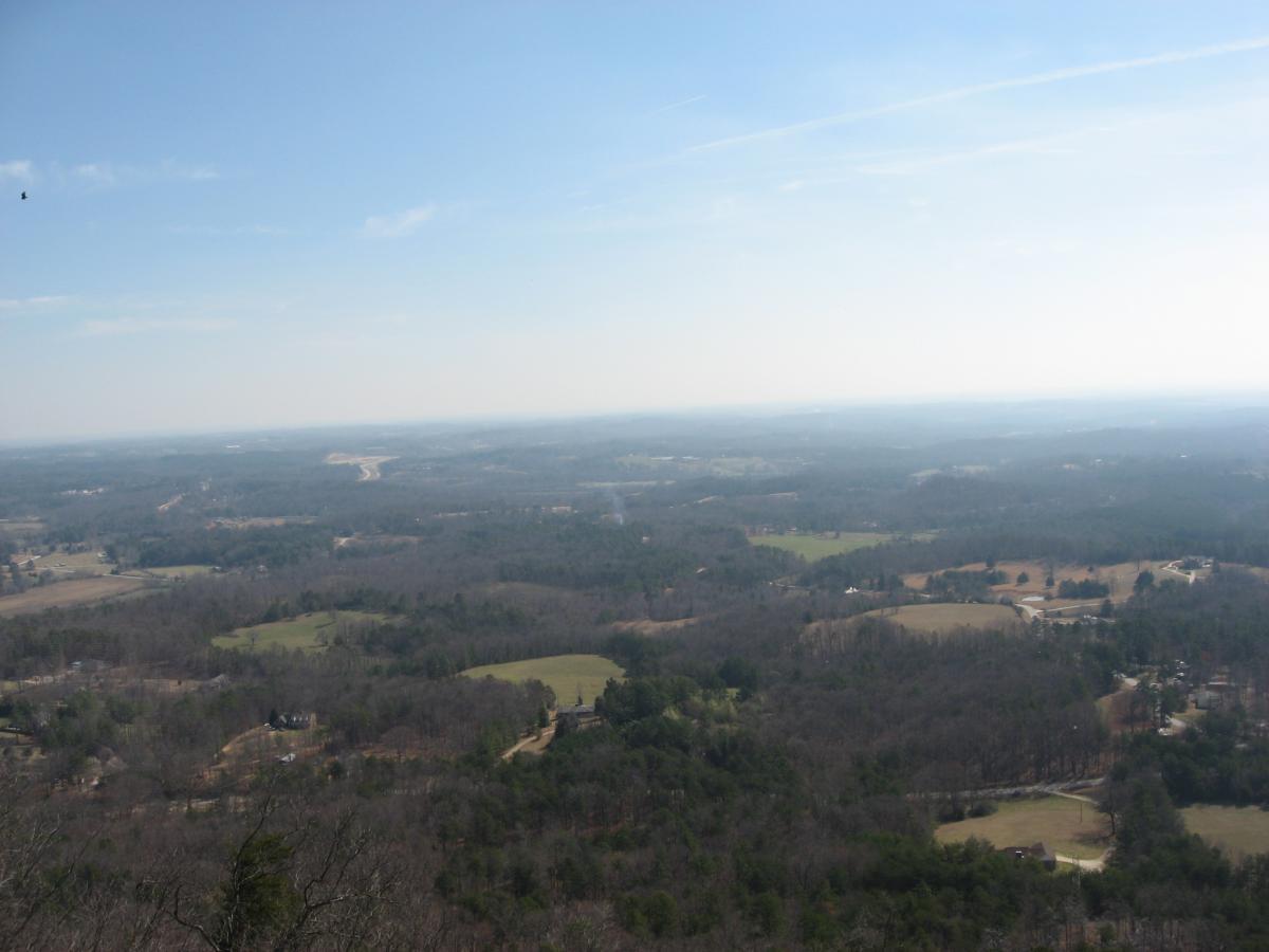 A panoramic view of a lush green landscape with rolling hills and far-off horizons under a clear blue sky. The scene includes patches of farmland and small clusters of trees, showcasing a serene natural environment. Currahee Mountain Road mountain bike trail.