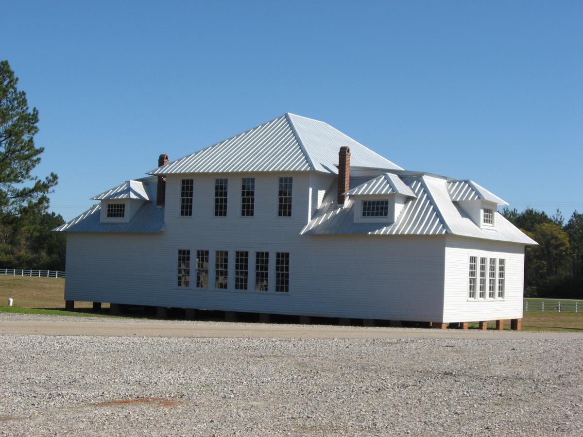A large, white wooden house with a metal roof, featuring multiple gabled sections and large windows, standing elevated on posts in a grassy field. The sky is clear and blue, with trees in the background and a gravel foreground. Heritage Park mountain bike trail.