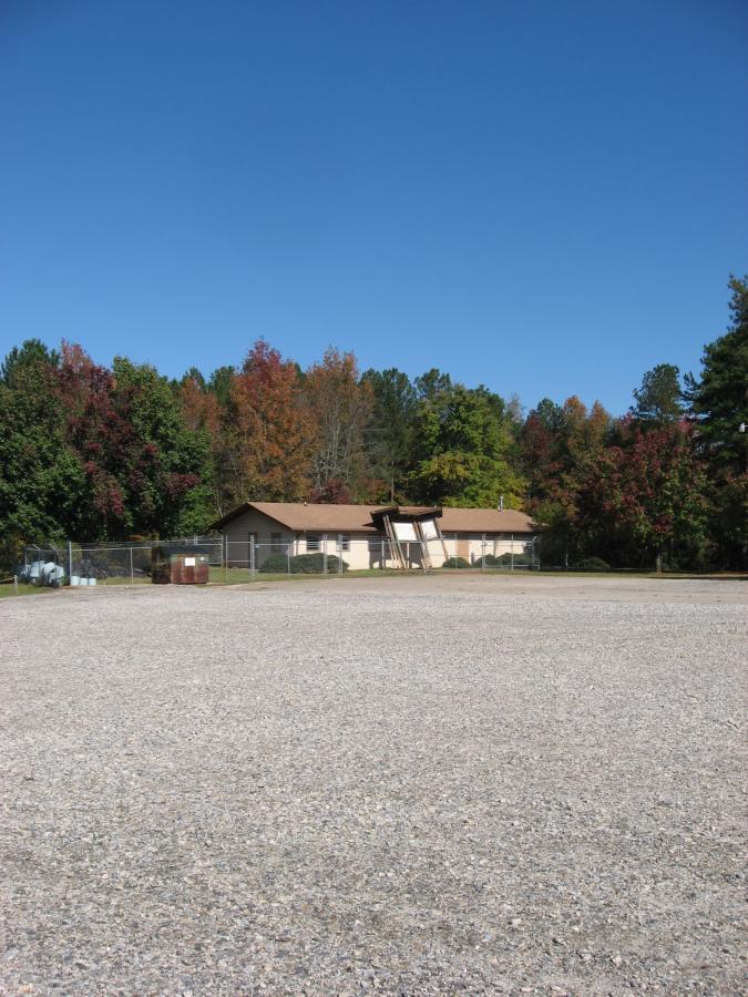 A view of a gravel lot in the foreground with a fenced building in the background, surrounded by trees in varying autumn colors under a clear blue sky. Heritage Park mountain bike trail.
