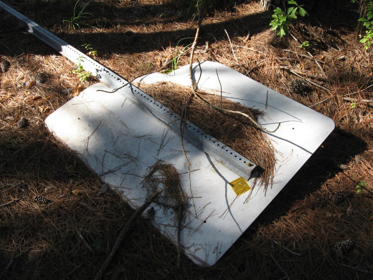 A rectangular metal sign resting on the forest floor, surrounded by pine needles and scattered pine cones. A thin branch and some dried grass are placed on top of the sign, with a metal mounting bracket visible on one side. Sunlight filters through the trees, creating dappled shadows. Jennings Mill mountain bike trail.