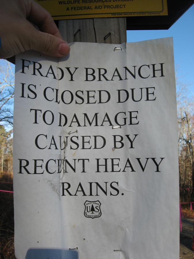 A sign pinned to a wooden post reading "Frady Branch is closed due to damage caused by recent heavy rains." The sign is partially wrinkled and has visible wear. In the background, trees and a blue sky can be seen. Frady Branch mountain bike trail.
