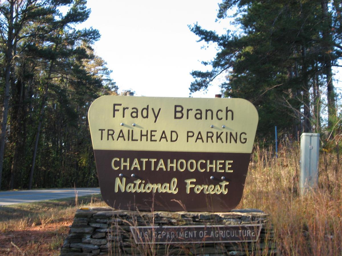 Sign for Frady Branch Trailhead Parking in Chattahoochee National Forest, with wooded surroundings and a paved road in the background. Frady Branch mountain bike trail.