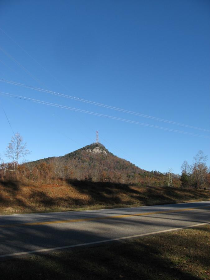 A hill topped with a communications tower is visible against a clear blue sky, with a rural road running in the foreground. The landscape features trees in autumn colors, indicating a seasonal change. Frady Branch mountain bike trail.