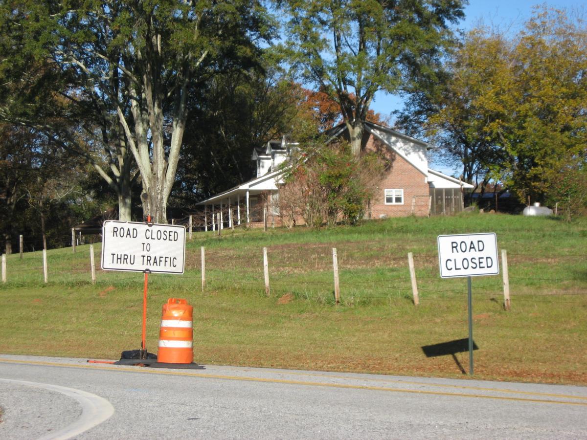 Two road closure signs are positioned along a curved road. One sign reads "ROAD CLOSED TO THRU TRAFFIC" and is accompanied by an orange construction barrel. The second sign, placed nearby, simply states "ROAD CLOSED." In the background, a grassy area and a house can be seen, along with several trees. Currahee Mountain Road mountain bike trail.
