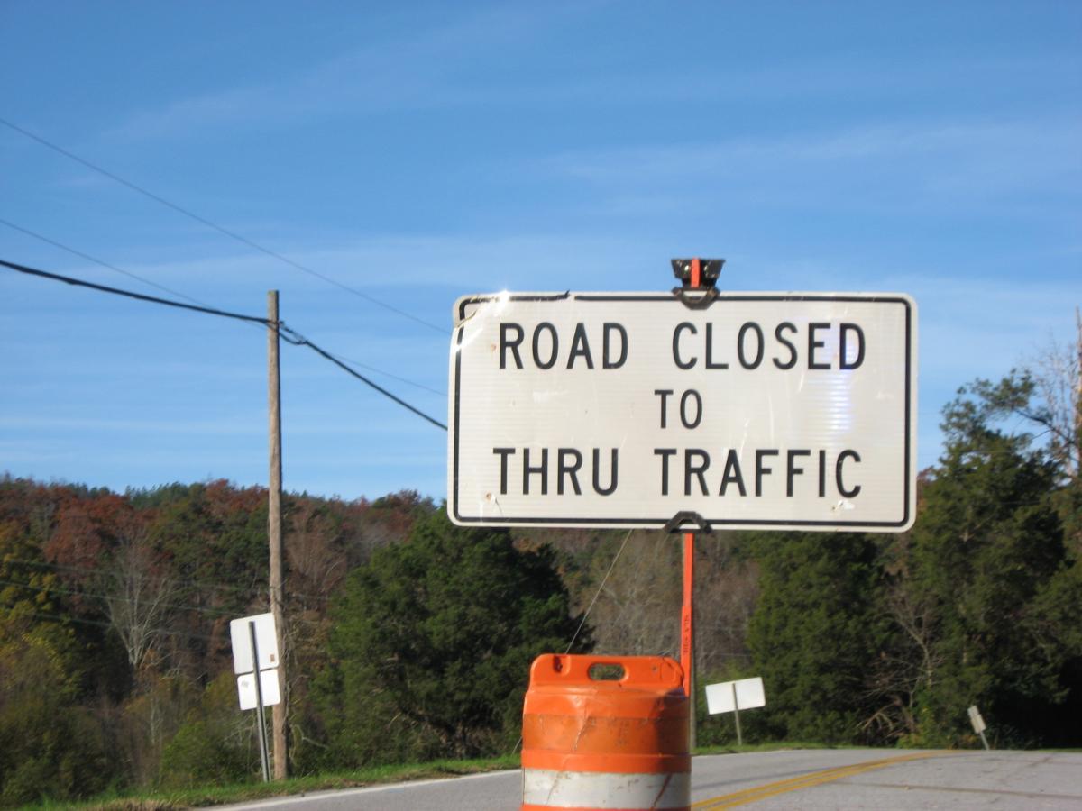 A road sign indicating "Road Closed to Thru Traffic," positioned near a barricade. The background features trees with autumn foliage and a clear blue sky. Currahee Mountain Road mountain bike trail.