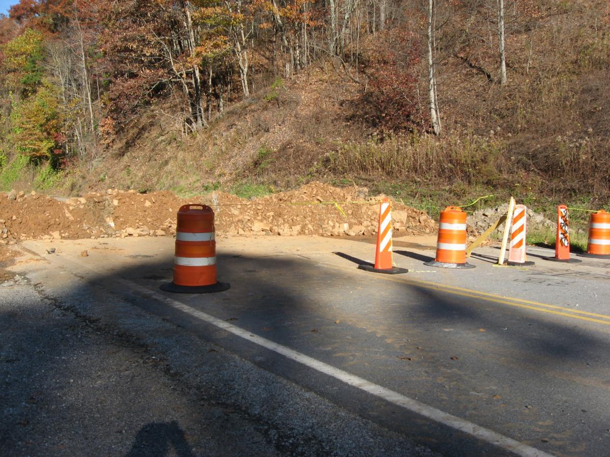 A view of a road under construction, featuring orange traffic cones and a barricade of dirt blocking the way. There are various traffic control devices indicating road closure, with trees and autumn foliage in the background. Currahee Mountain Road mountain bike trail.