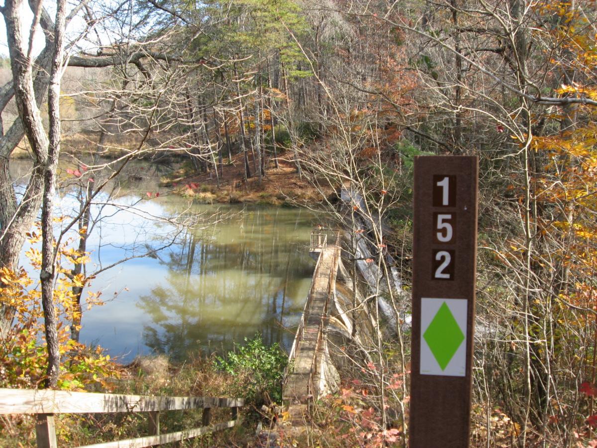 A serene view of a tranquil lake surrounded by trees in autumn foliage. In the foreground, a wooden sign displays the numbers "152" and a green diamond symbol, indicating a trail marker. The water reflects the surrounding landscape, and a wooden bridge extends over the lake, leading to a small dock. Lake Russell Loop mountain bike trail.