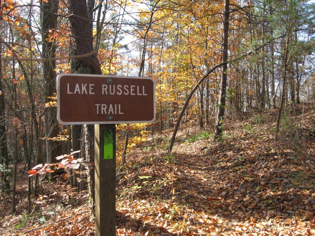 A brown wooden sign reads "Lake Russell Trail," indicating the beginning of a hiking path surrounded by trees with colorful autumn leaves. The trail is covered in fallen leaves, and the sun is shining through the branches, creating a serene outdoor atmosphere. Lake Russell Loop mountain bike trail.