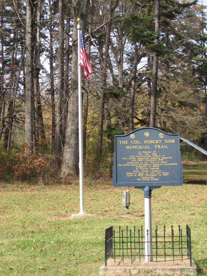 A memorial plaque for Colonel Robert Sink, located on a trail surrounded by trees. An American flag is flying on a pole nearby. The plaque honors Colonel Sink, highlighting his service and contributions, with inscriptions detailing his military history. The setting features a peaceful, wooded area. Currahee Mountain Road mountain bike trail.
