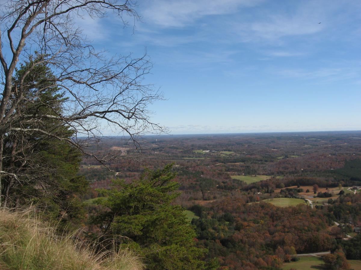 A scenic view from a high vantage point, featuring a landscape of rolling hills and valleys. The foreground includes bare branches and evergreen trees, while the background displays a stretch of countryside with patches of colorful foliage in various autumn hues, under a clear blue sky. Currahee Mountain Road mountain bike trail.