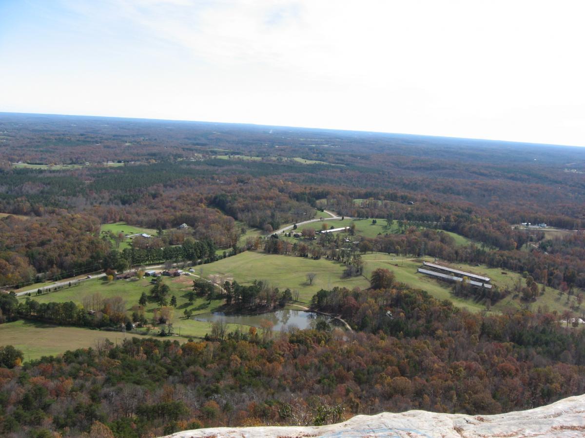 A panoramic view from a high vantage point, showcasing rolling hills and valleys covered in autumn foliage. The landscape features a mix of green fields, trees with changing leaves, a small pond, and several buildings along a winding road. The sky is partly cloudy, enhancing the scenic beauty of the countryside. Currahee Mountain Road mountain bike trail.