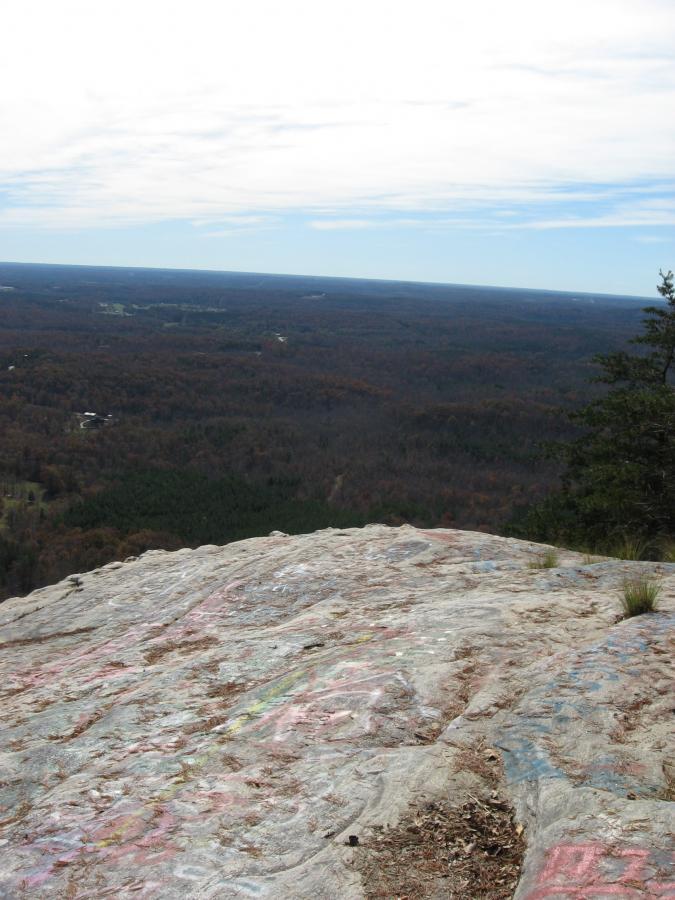 A panoramic view from a rocky cliff overlooking a forested landscape. The foreground features a large, smooth, and graffitied rock surface, while the background displays rolling hills covered in autumn foliage, ranging from shades of green to orange and brown. The sky above is mostly cloudy with some visible blue, creating a serene atmosphere. Currahee Mountain Road mountain bike trail.