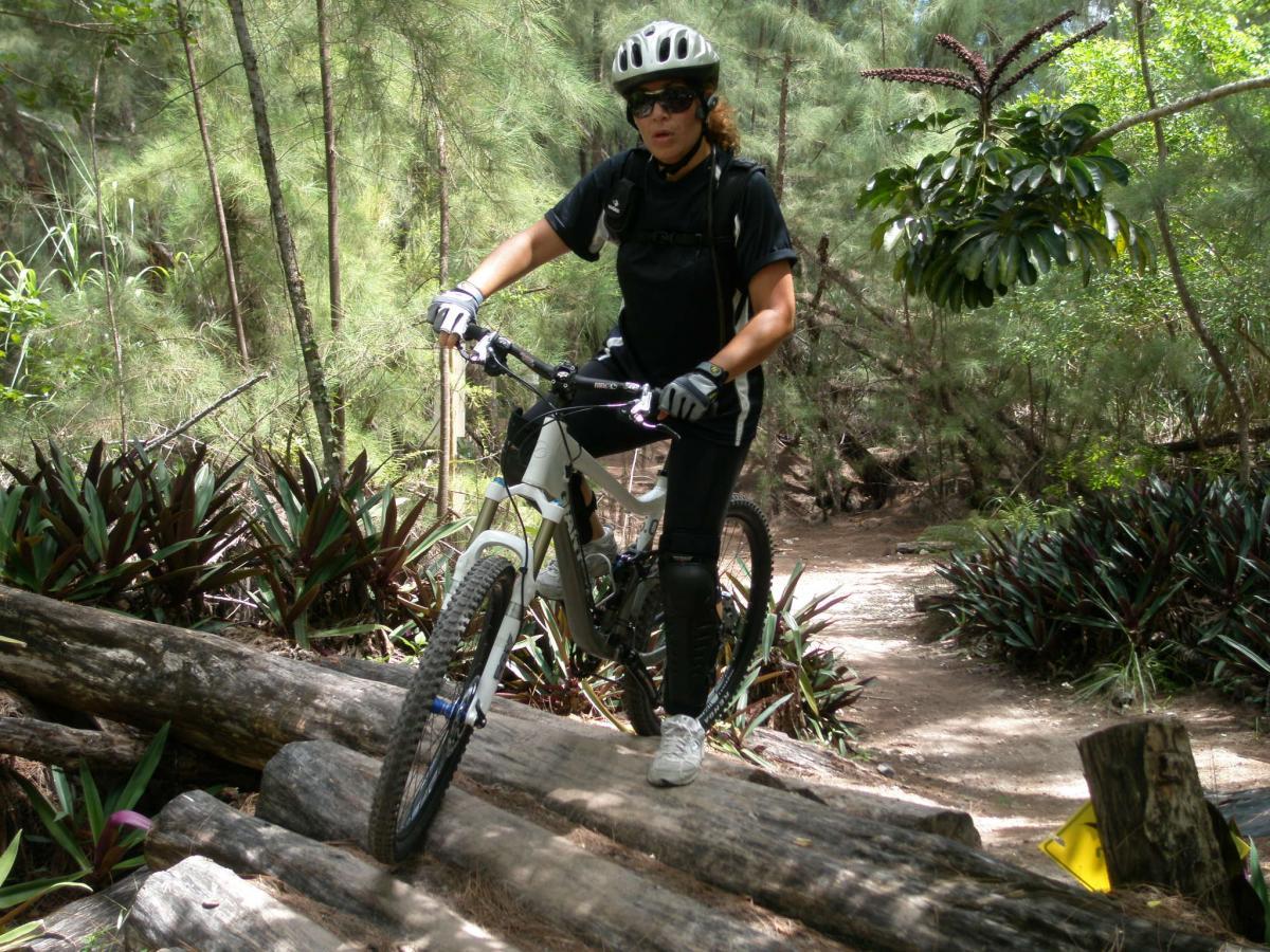 A woman with curly hair wearing a helmet and sunglasses is navigating a mountain bike over a series of wooden logs on a trail surrounded by lush greenery. She is dressed in a black cycling outfit and is focused on maintaining balance as she rides through the natural landscape. Oleta River State Park mountain bike trail.