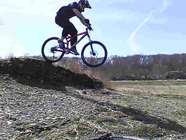 A person in a black helmet and clothing is riding a mountain bike off a dirt ramp, capturing a moment of mid-air action. The background features an open field and trees under a clear blue sky. Great Esker Park mountain bike trail.