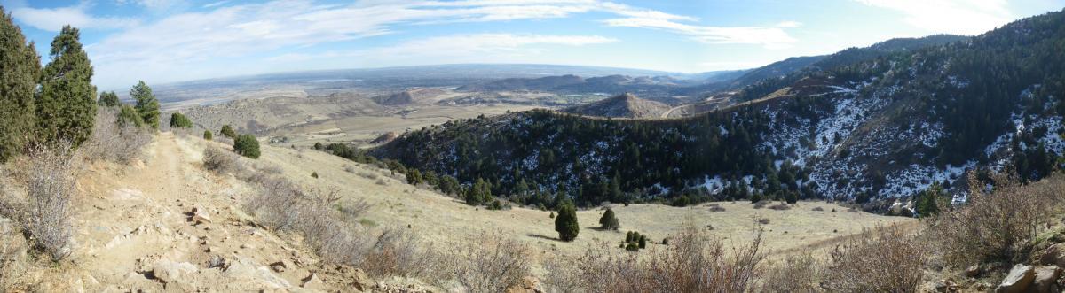 A panoramic view of a mountainous landscape, featuring rolling hills and valleys under a clear blue sky. The foreground includes a rocky trail with sparse vegetation and patches of snow. In the background, the terrain transitions from rocky outcrops to green hills, extending to a distant horizon that showcases the scenic beauty of the natural area. Mount Falcon Park mountain bike trail.