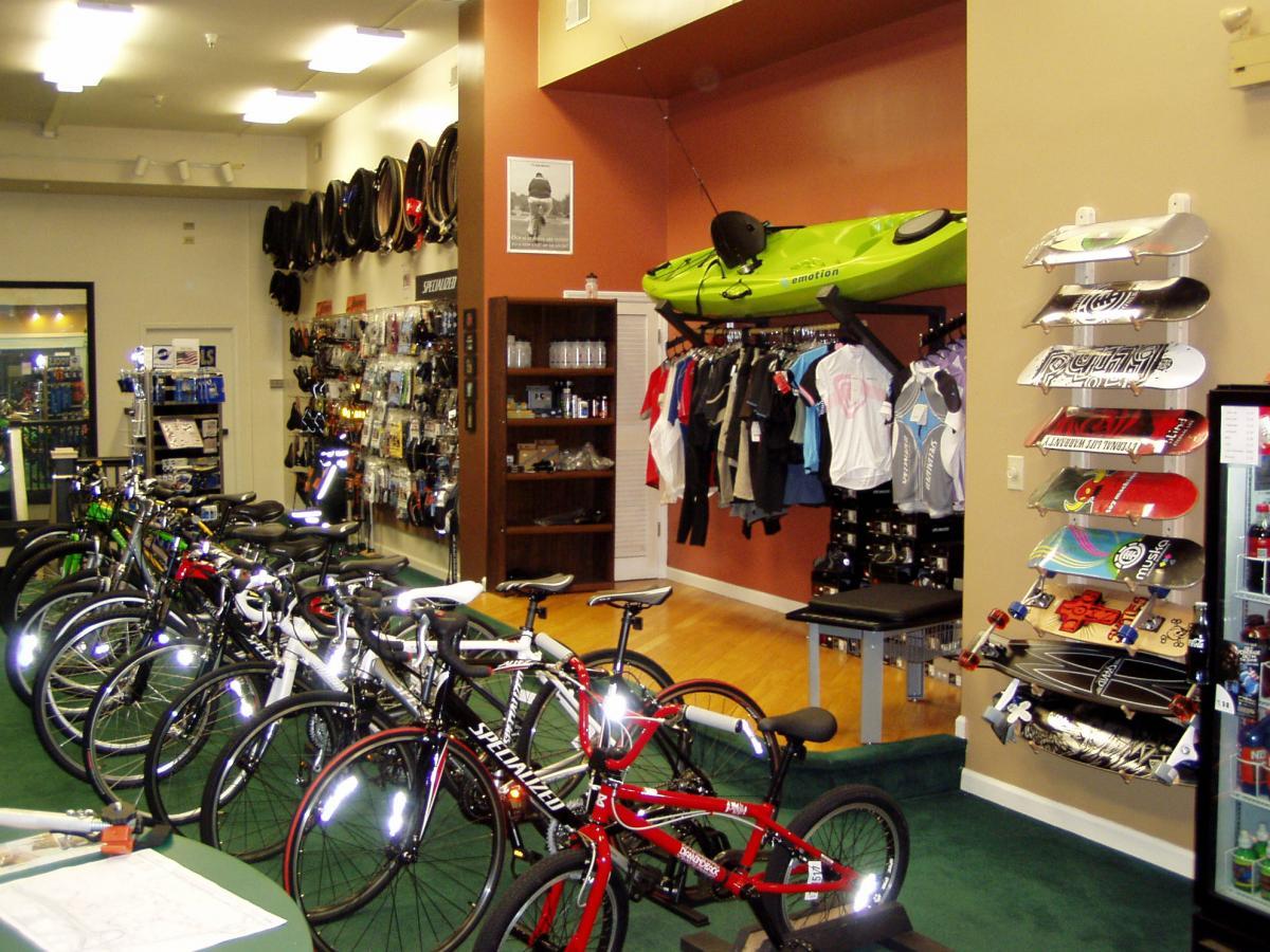Alt text: An indoor sporting goods store featuring a row of colorful bicycles, various biking accessories displayed on shelves, a green kayak mounted on the wall, and skateboards hanging on racks. The store has a bright and organized layout with a green carpeted floor and several clothing items showcased.
