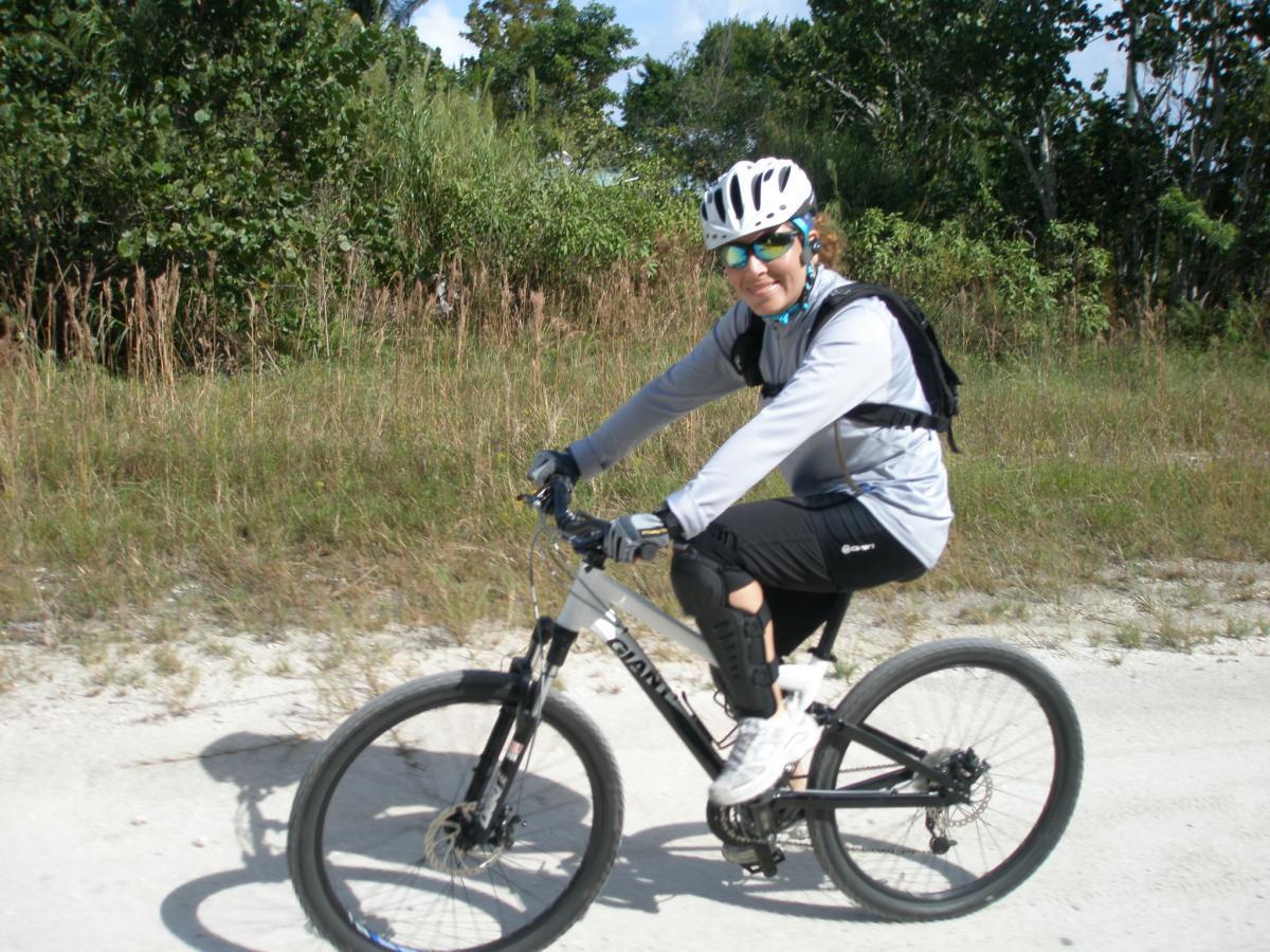 A person wearing a helmet and sunglasses rides a mountain bike along a dirt path surrounded by greenery. They are dressed in athletic gear, including a long-sleeve shirt and pants, suitable for biking. The sun is shining, creating a bright and active outdoor atmosphere. Oleta River State Park mountain bike trail.