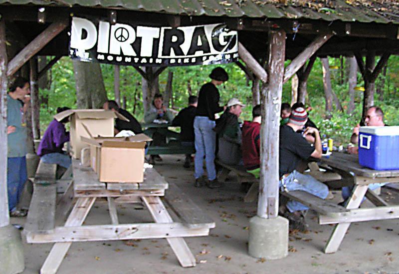 A group of people gathered under a wooden pavilion, participating in a social event. A banner reading "DIRTRAG" hangs above, indicating a connection to mountain biking. Several individuals are seated at picnic tables, with some interacting while others appear to be preparing food or drinks. The setting is surrounded by trees, suggesting an outdoor location. North Park mountain bike trail.