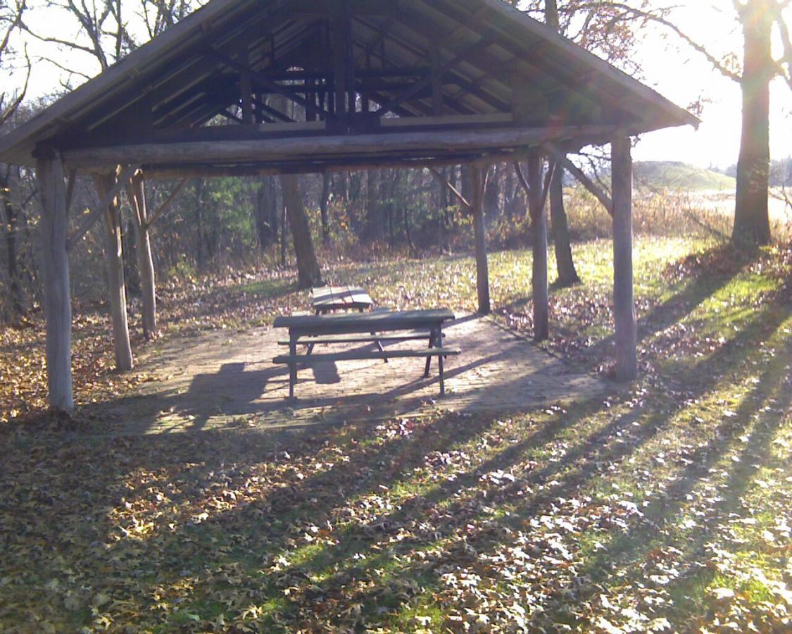 A rustic pavilion in a wooded area, featuring a wooden picnic table underneath. The ground is covered with fallen leaves, and long shadows are cast across the area by the low sun. Trees surround the space, providing a natural and serene atmosphere. Heritage Park mountain bike trail.