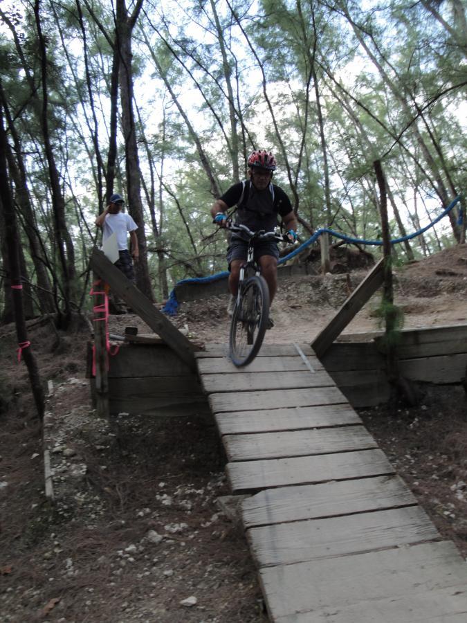 A mountain biker in a helmet and protective gear is riding down a wooden ramp during a trail ride in a forested area. A person can be seen in the background observing. The scene is surrounded by tall trees and natural vegetation. Oleta River State Park mountain bike trail.