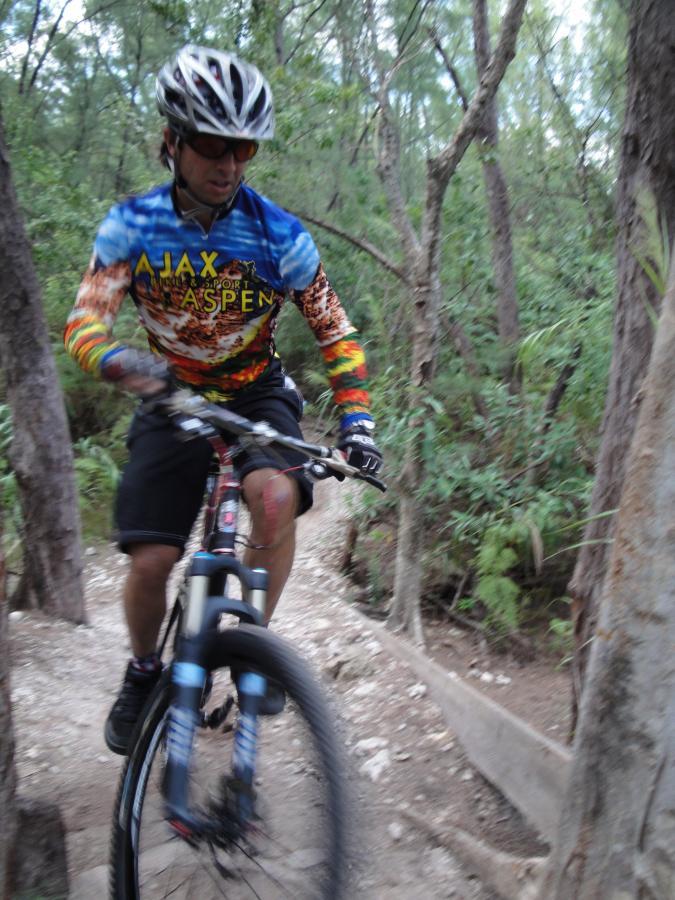 A mountain biker wearing a colorful long-sleeve jersey and protective gear rides down a narrow trail surrounded by trees and greenery. The image captures the dynamic motion of the rider navigating the terrain. Oleta River State Park mountain bike trail.
