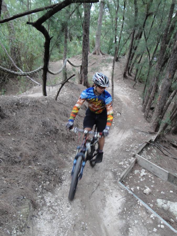 A mountain biker wearing a colorful tie-dye shirt and helmet navigates a winding dirt trail surrounded by trees. The setting features a mix of gravel and dirt paths under a canopy of foliage. Oleta River State Park mountain bike trail.