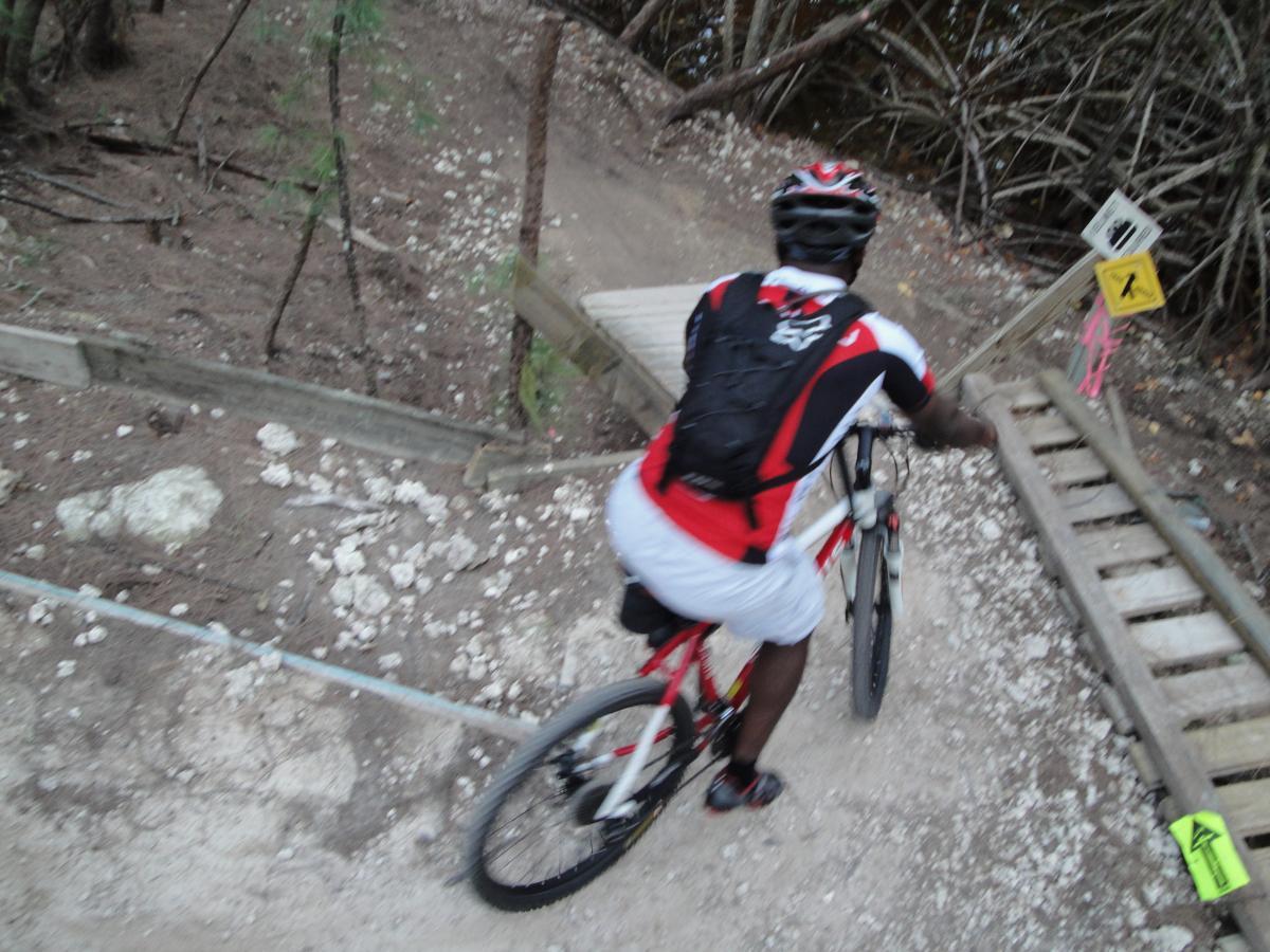 A cyclist riding a mountain bike on a narrow dirt trail, approaching a wooden bridge. The surrounding area features rocky terrain and sparse vegetation, with caution signs indicating potential hazards ahead. The cyclist is wearing a helmet, a black backpack, and a red-and-white cycling outfit. Oleta River State Park mountain bike trail.