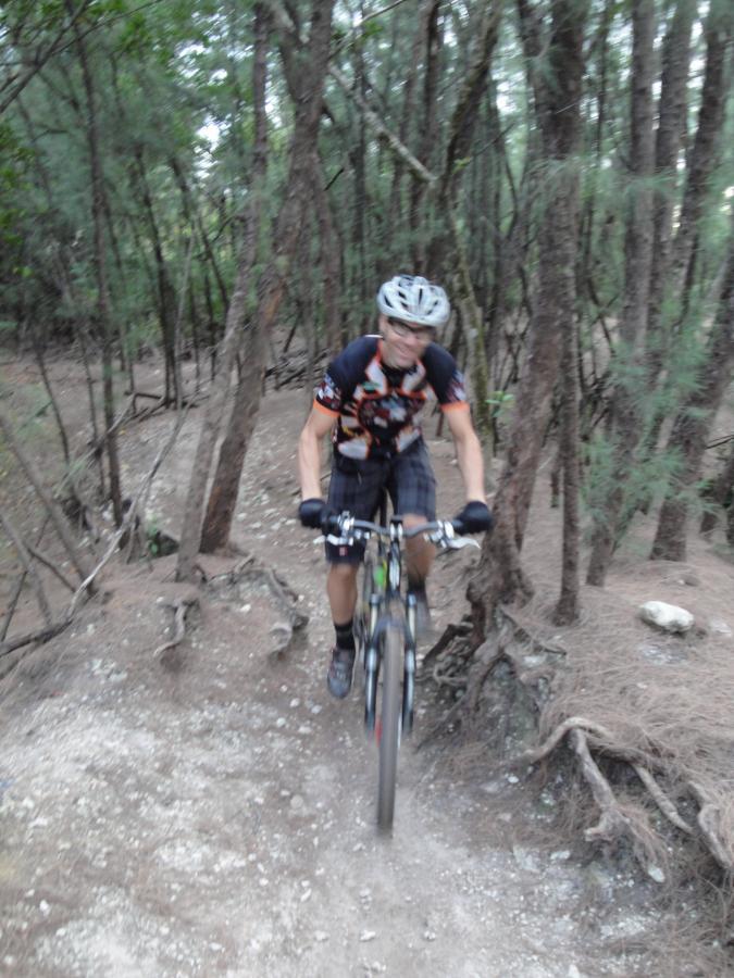 A mountain biker riding on a dirt trail surrounded by trees, wearing a helmet and cycling gear. The bike is slightly lifted off the ground, suggesting motion and speed as the cyclist navigates the rugged terrain. Oleta River State Park mountain bike trail.
