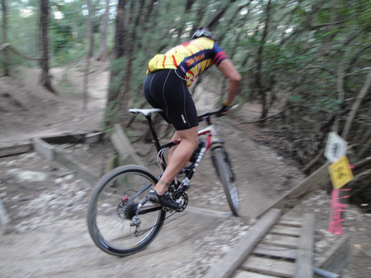 A cyclist in a colorful jersey performs a jump on a mountain bike over a wooden bridge on a dirt trail, surrounded by trees in a wooded area. The image captures the motion and excitement of mountain biking. Oleta River State Park mountain bike trail.