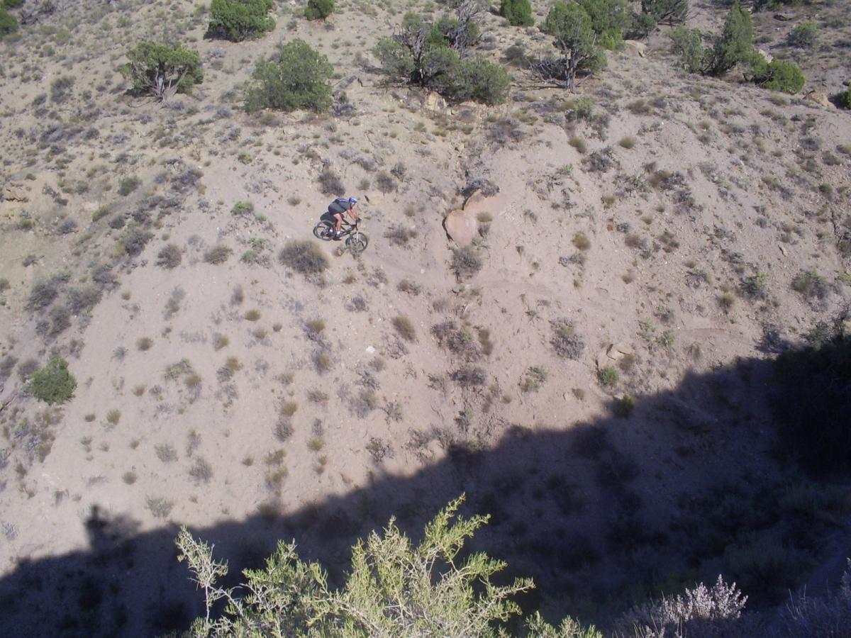 A mountain biker navigating a rugged, sandy hillside surrounded by sparse vegetation and scattered rocks under a clear sky. Palisade Rim mountain bike trail.