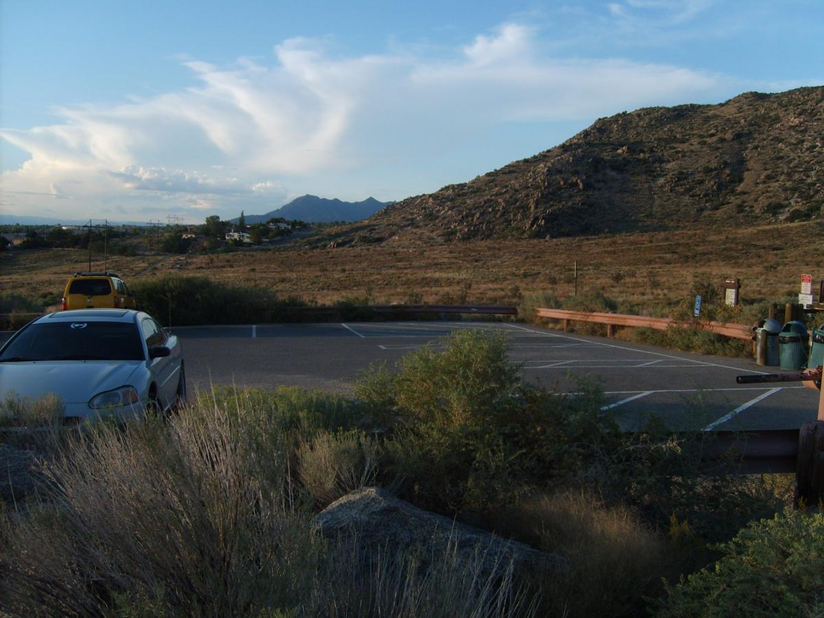A parking lot surrounded by sparse vegetation, with a few cars parked, notably a silver sedan in the foreground and a yellow vehicle in the background. The landscape features hills and mountains in the distance under a partly cloudy sky, suggesting a natural setting. Sandia Mountains Foothill Trail mountain bike trail.