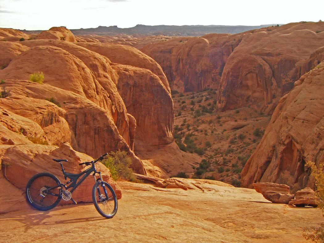 A mountain bike resting on rocky terrain with a canyon view in the background, showcasing red rock formations and sparse vegetation under a bright sky. Slickrock mountain bike trail.