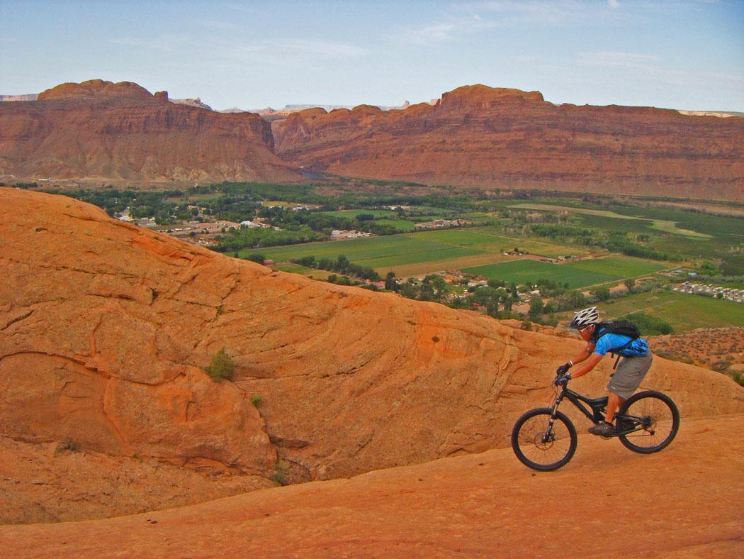 A mountain biker riding on a rocky, red terrain with a scenic view of a green valley and distant mountains in the background. The sky is mostly clear with a few clouds, highlighting the adventurous outdoor setting. Slickrock mountain bike trail.
